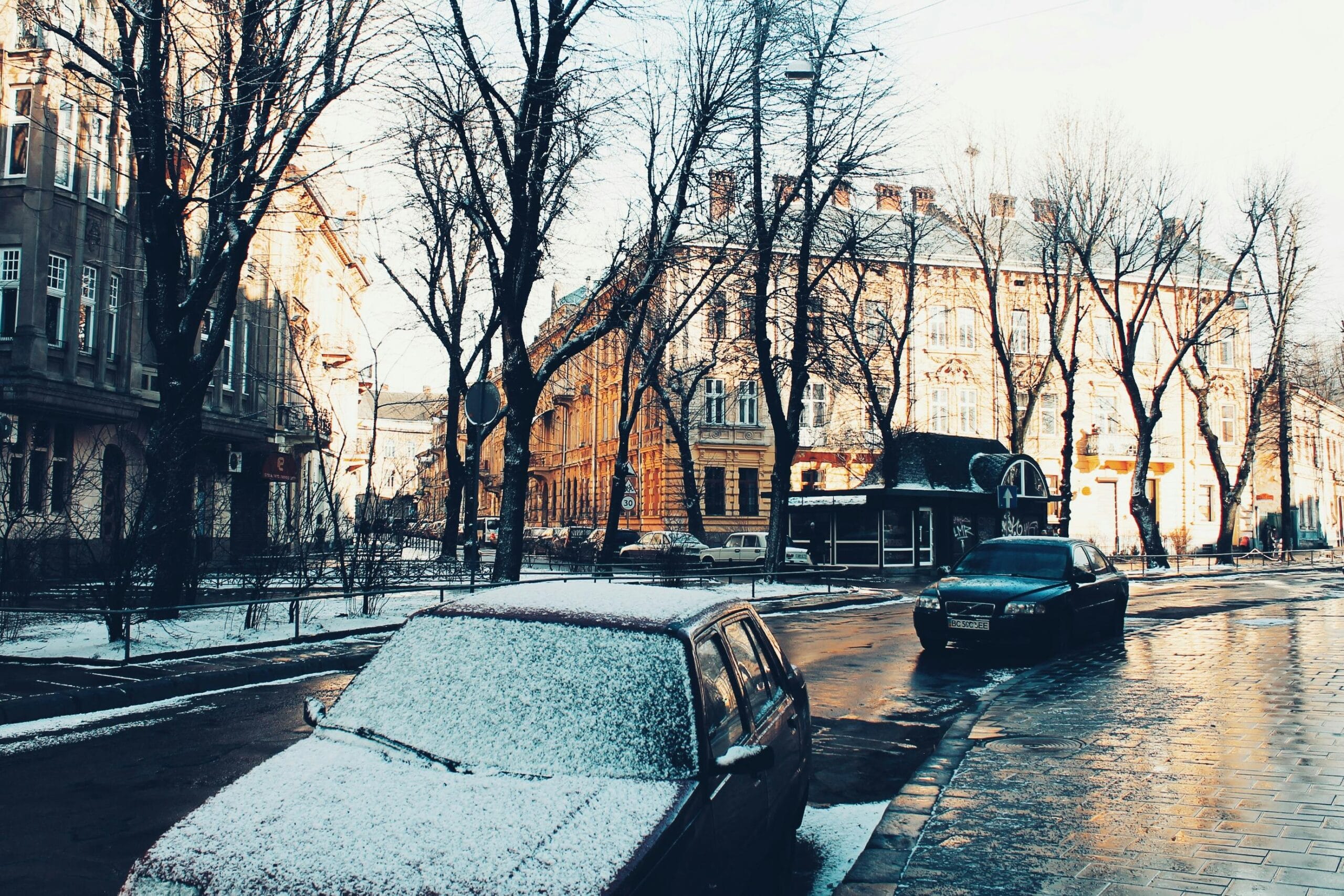 Snow-covered car parked on a city street lined with bare trees and classical buildings in winter sunlight