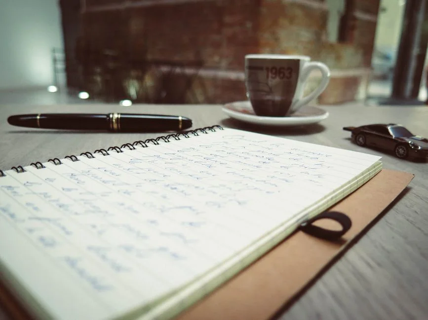 A spiral notepad and pen beside a coffee cup on a desk