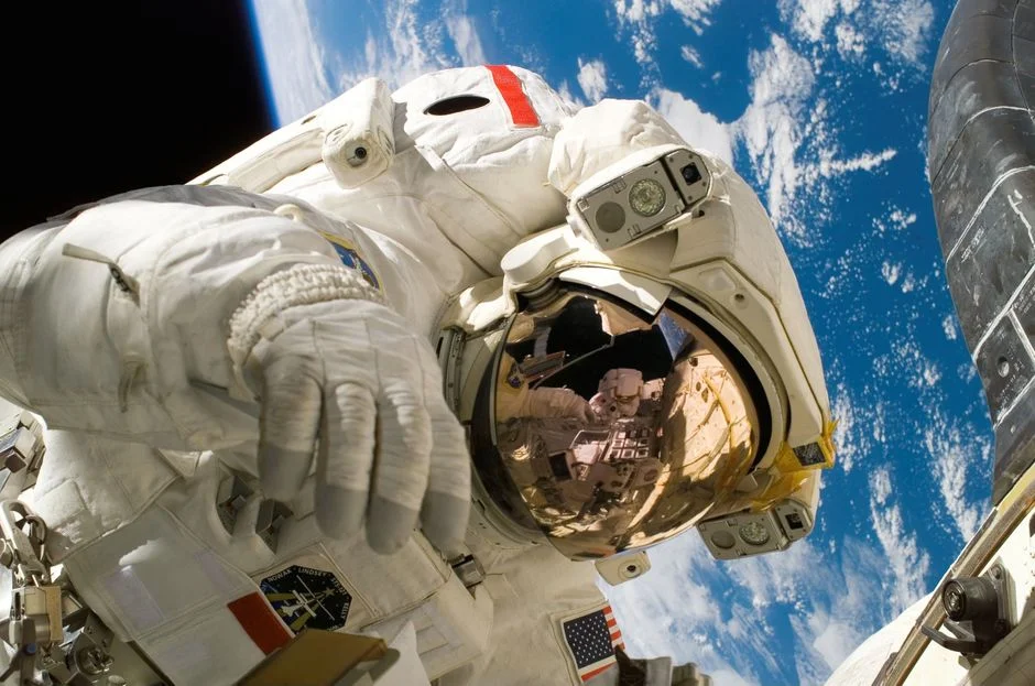 Astronaut in a white spacesuit floating outside a spacecraft with Earth and clouds behind, reflected in the gold helmet visor