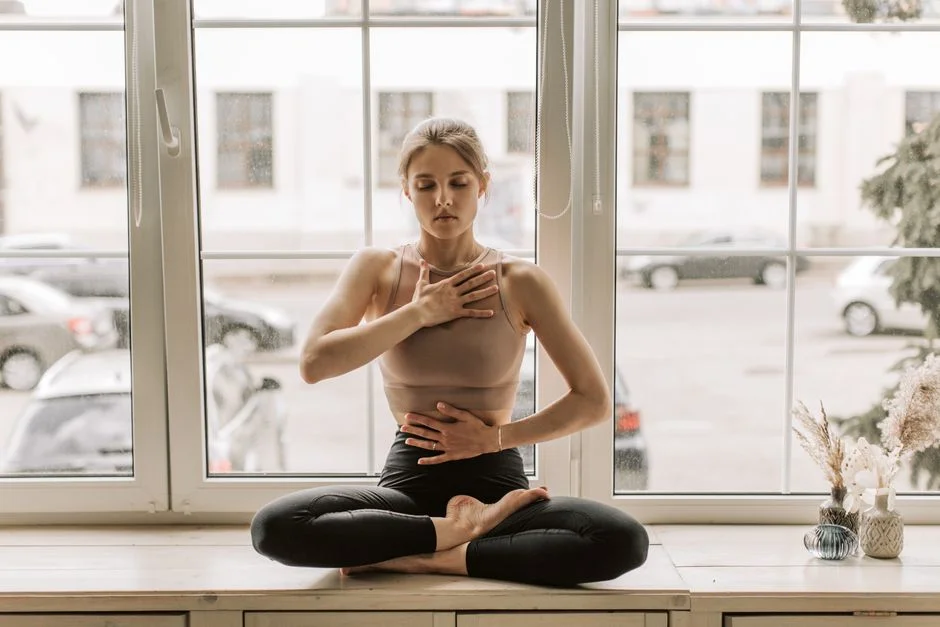 Woman sitting cross-legged on a window ledge with one hand on her chest and one on her belly, eyes closed, feeling her breath