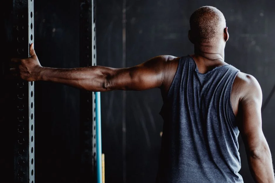 Man seen from behind stretching one arm out to the side against a gym rack to open his chest and shoulder