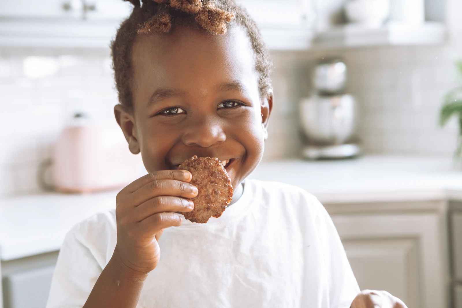 Smiling child in a white shirt biting into a brown round snack in a bright kitchen