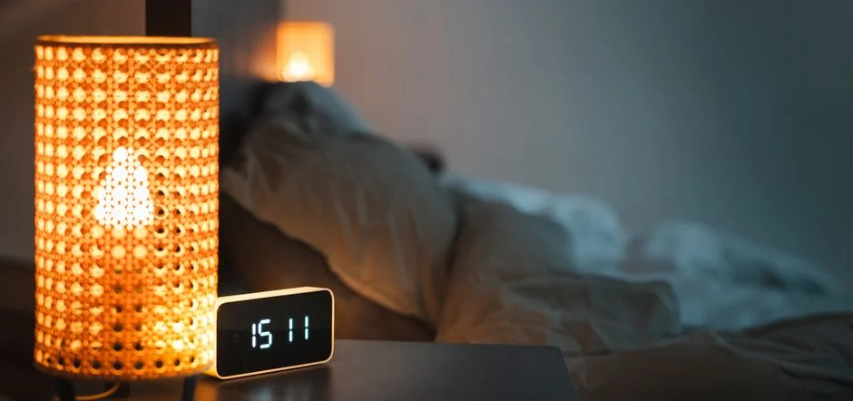 Dim bedroom at night with a glowing woven lamp and a digital clock on the nightstand beside an unmade bed
