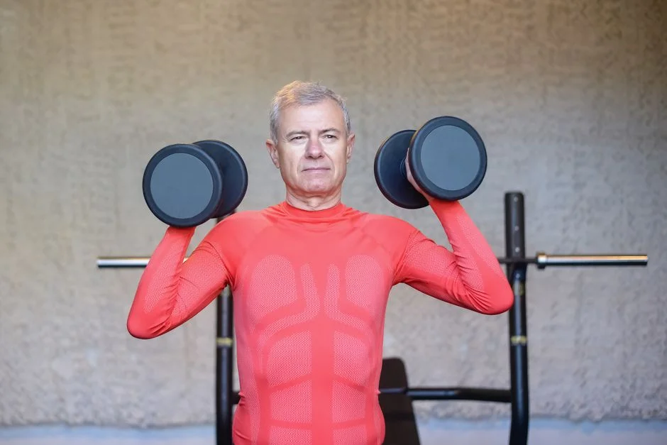 Older man with gray hair holding two dumbbells at shoulder height during a strength workout