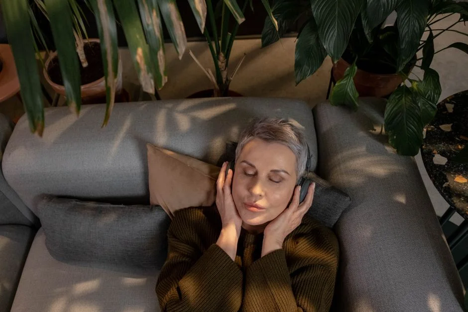 Woman with short gray hair lying on a sofa with her eyes closed, holding headphones to her ears