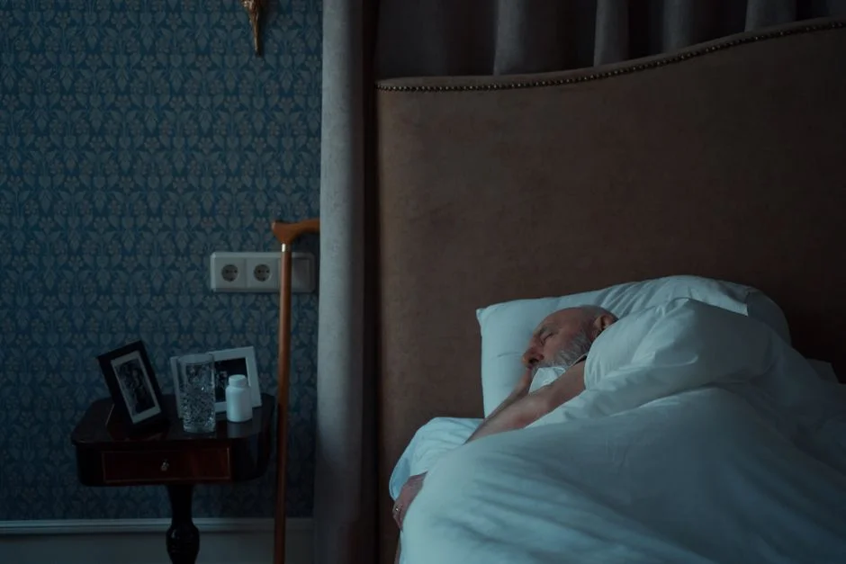 Older man with a gray beard asleep under a white duvet in a dark bedroom