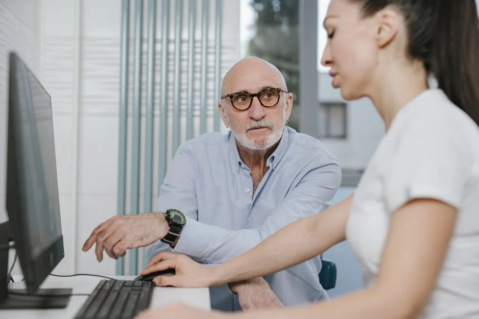 Concerned older man in glasses talking with a clinician at a desk computer during a consultation