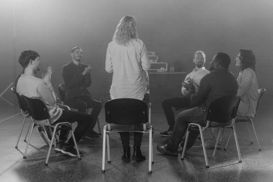 Adults seated in a circle of chairs as one person stands to speak while others listen and applaud