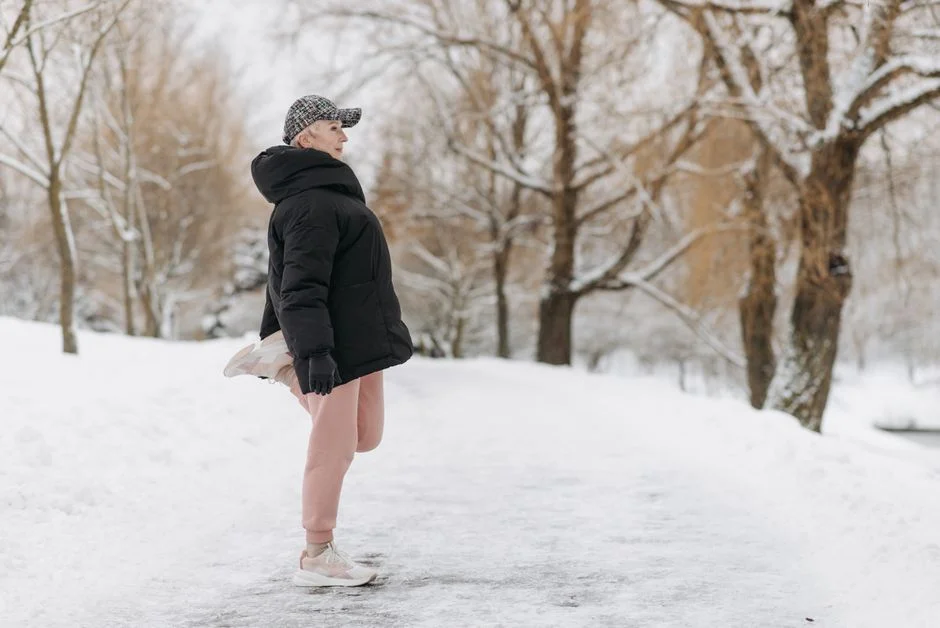 Older woman in a puffer jacket stretching her leg on a snowy park path