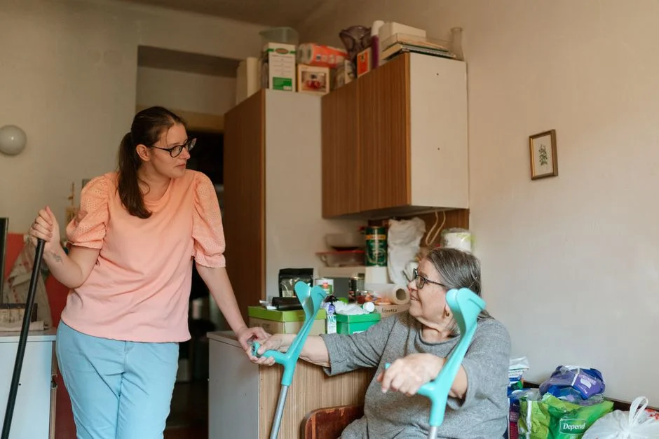 Younger woman talking with a seated older woman who has crutches, in a home kitchen
