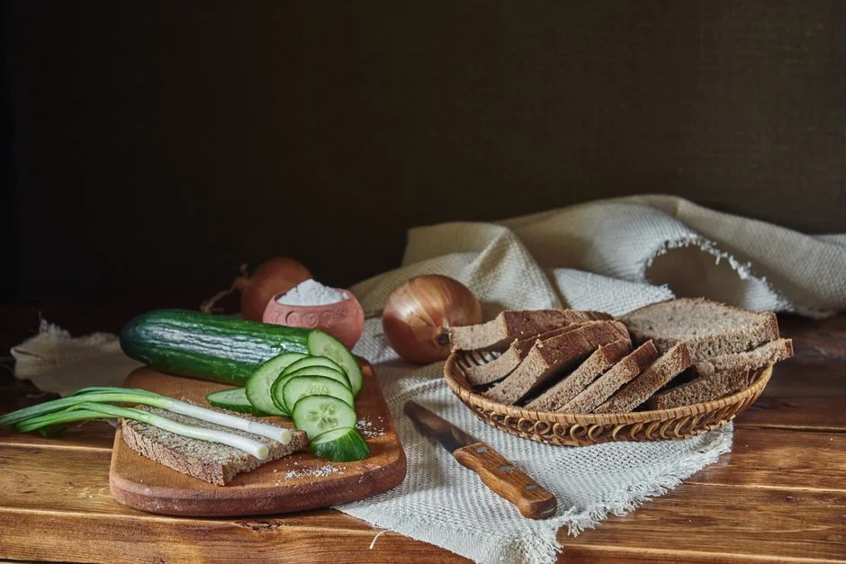 Slices of dark rye bread in a wicker basket beside cucumber and green onions on a wooden table