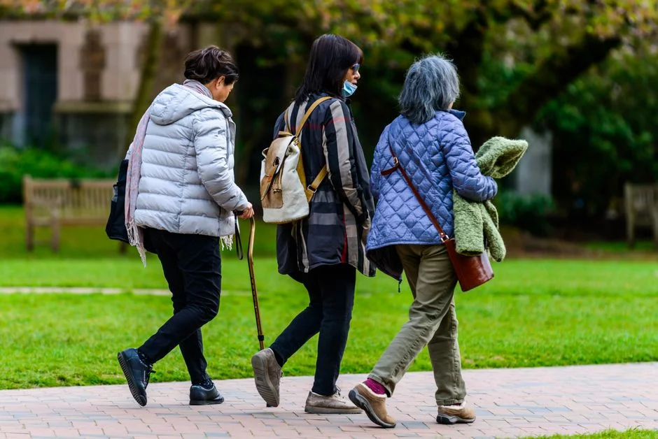 Three older women walking together along a paved path beside a lawn, one using a cane