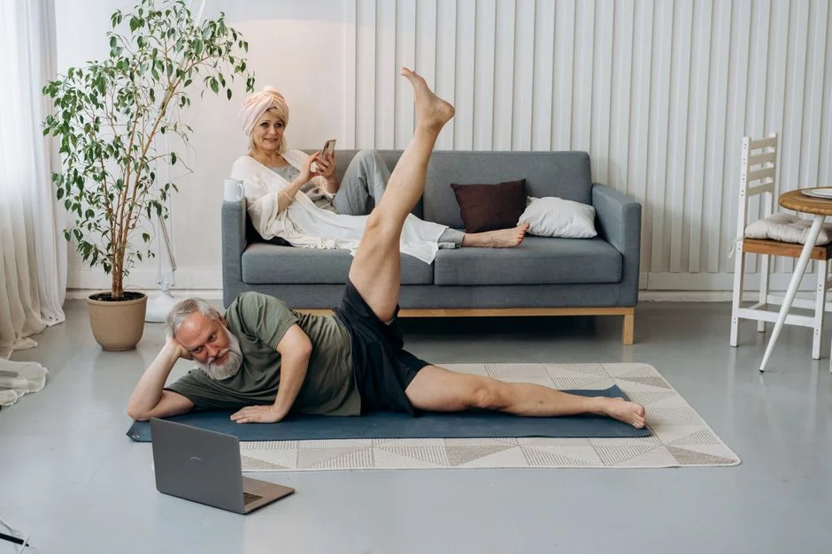 Older man doing a side-lying leg raise on a yoga mat in a living room while following a laptop