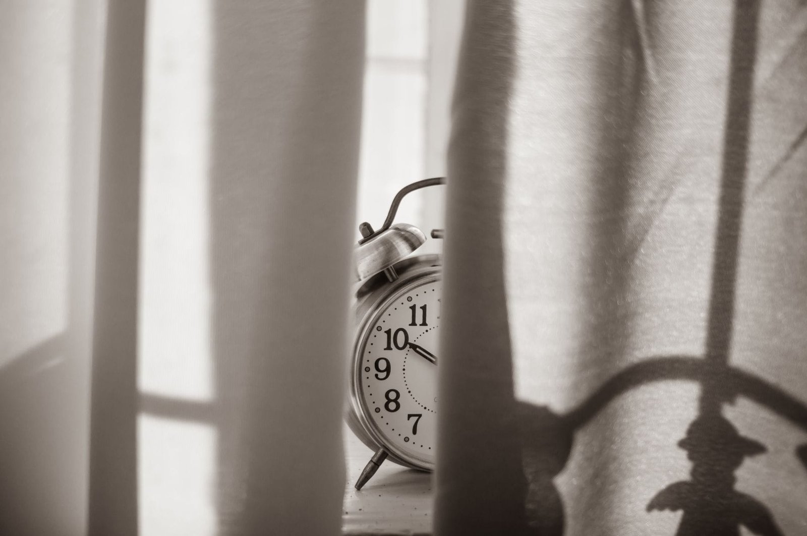Vintage alarm clock beside a sunlit window curtain in black and white
