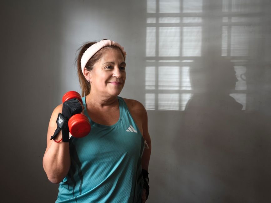 Smiling woman in a teal tank top and headband lifting a red dumbbell in a bare room